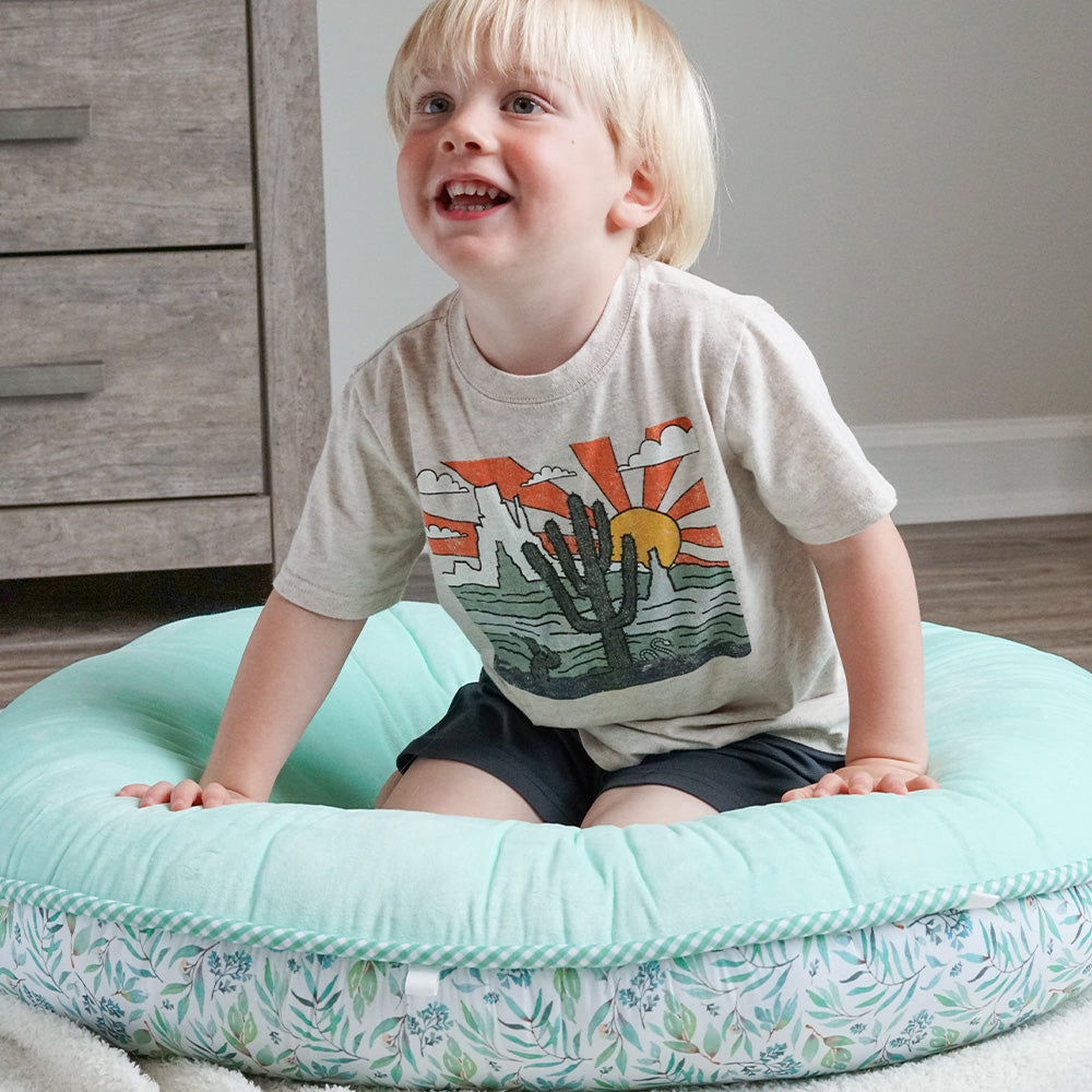Lifestyle picture of a young boy sitting and smiling on the Basil Green Pello Floor Cushion.