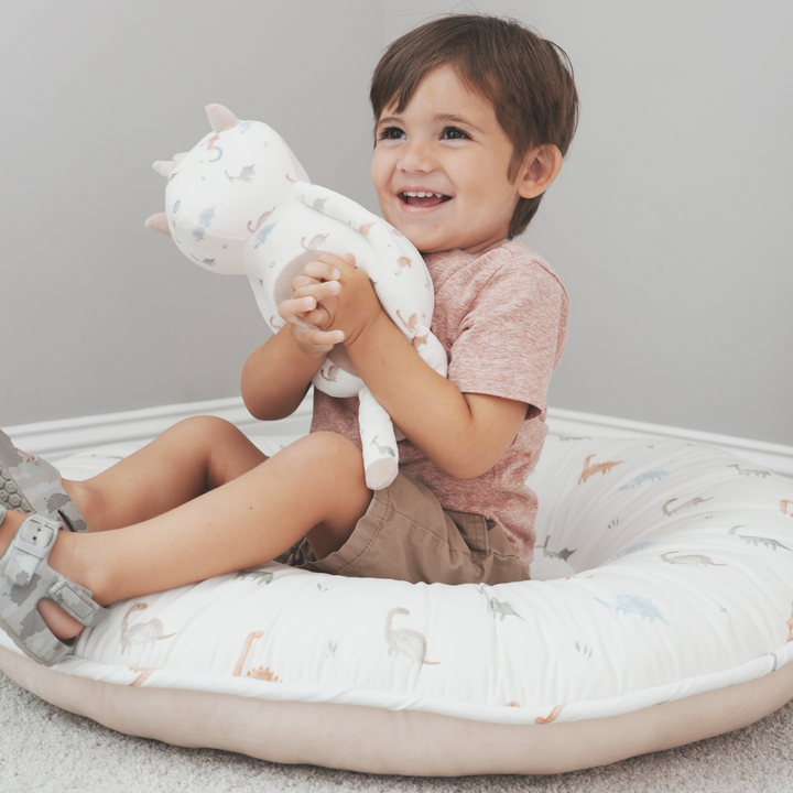 Darby Dino Floor Cushion with a smiling toddler boy sitting in it and holding Darby Dino plush.