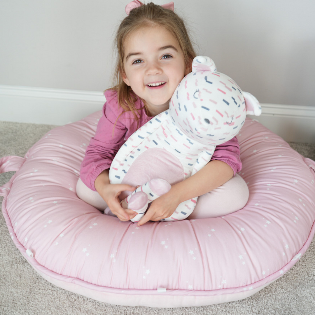 Pello Pink Estelle Floor Cushion with a smiling girl sitting in it holding a stuffed animal.