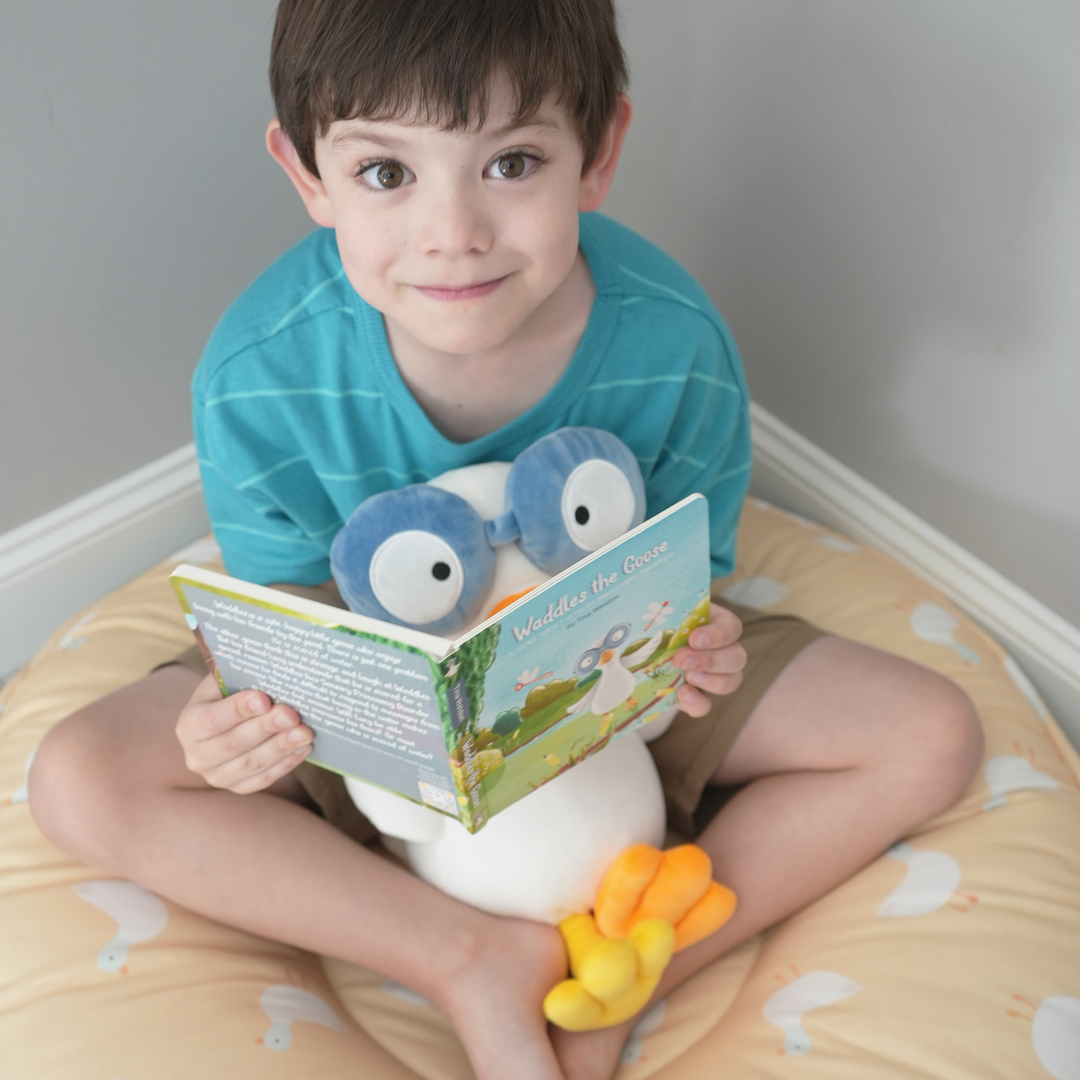 Puddles Whispering Blue Floor Cushion with a young boy sitting in it and reading Waddles the Goose board book while holding Waddles plush.