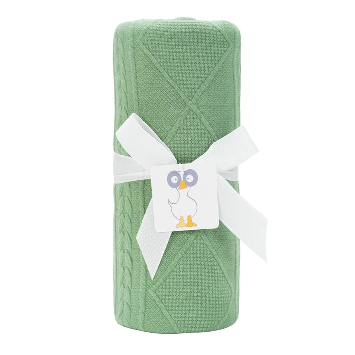 A sage green knit blanket with a cable pattern, wrapped with a white ribbon and tagged with a card featuring an owl.